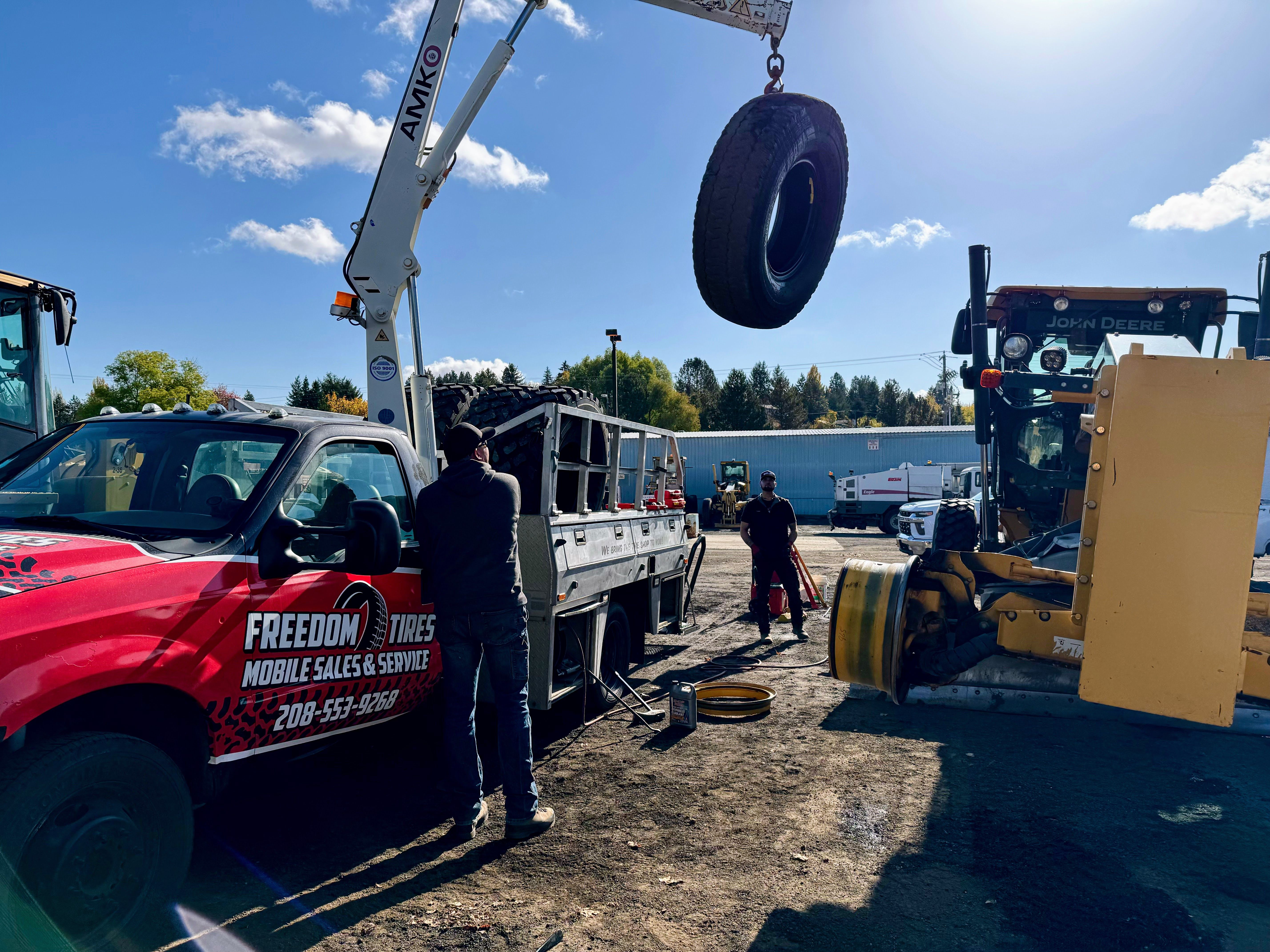 Freedom Tires crane lifting heavy tire at equipment yard with John Deere machinery